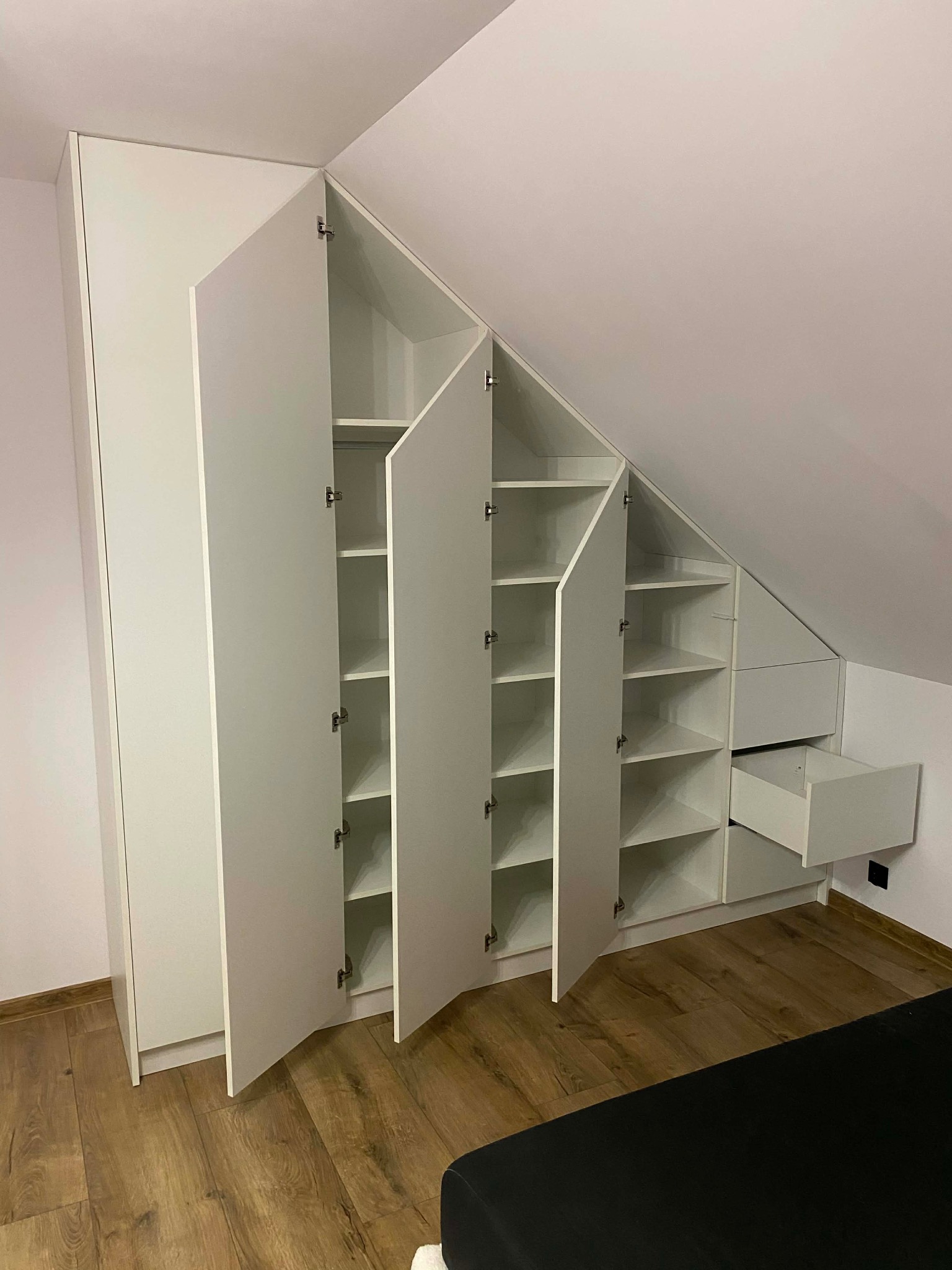 Biała szafa wnękowa na poddaszu ze skośnym sufitem, częściowo otwarta, ukazująca półki i szuflady. Widoczne drewniane panele podłogowe.
