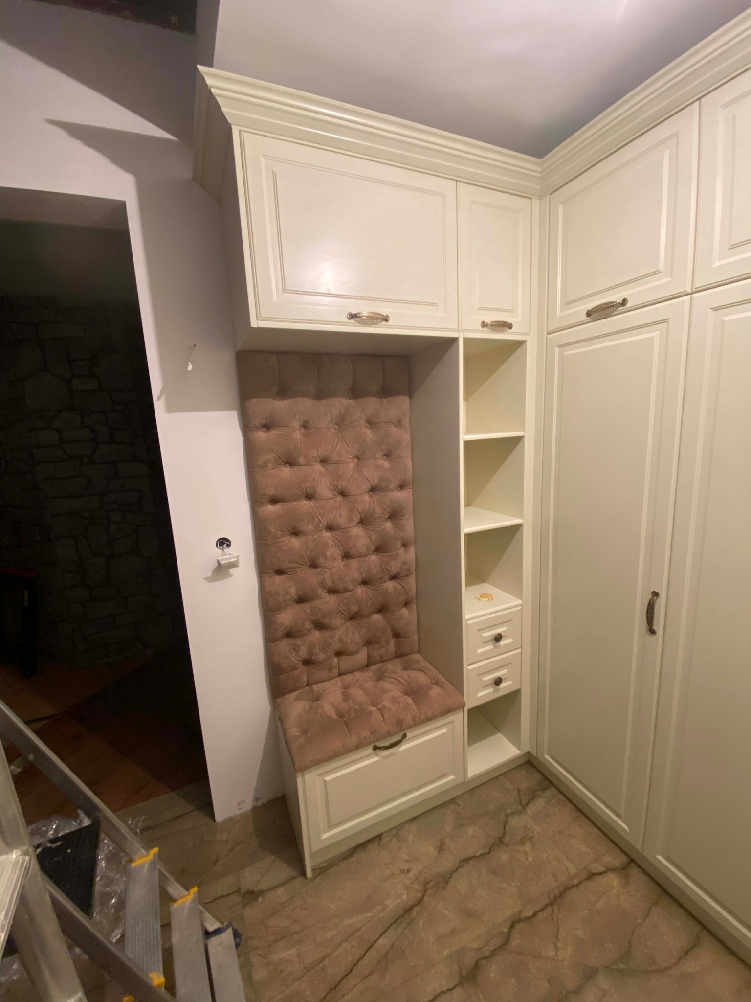 Biała szafa wnękowa z tapicerowanym siedziskiem i oparciem w kolorze pudrowego różu. Widoczne półki i szuflady. Podłoga z płytek imitujących marmur.