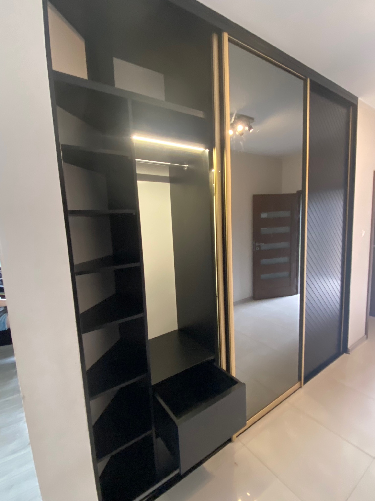 Czarna szafa wnękowa z przesuwnymi drzwiami lustrzanymi i otwartymi półkami, oświetlona listwą LED, widok na wnętrze pokoju z drewnianymi drzwiami.