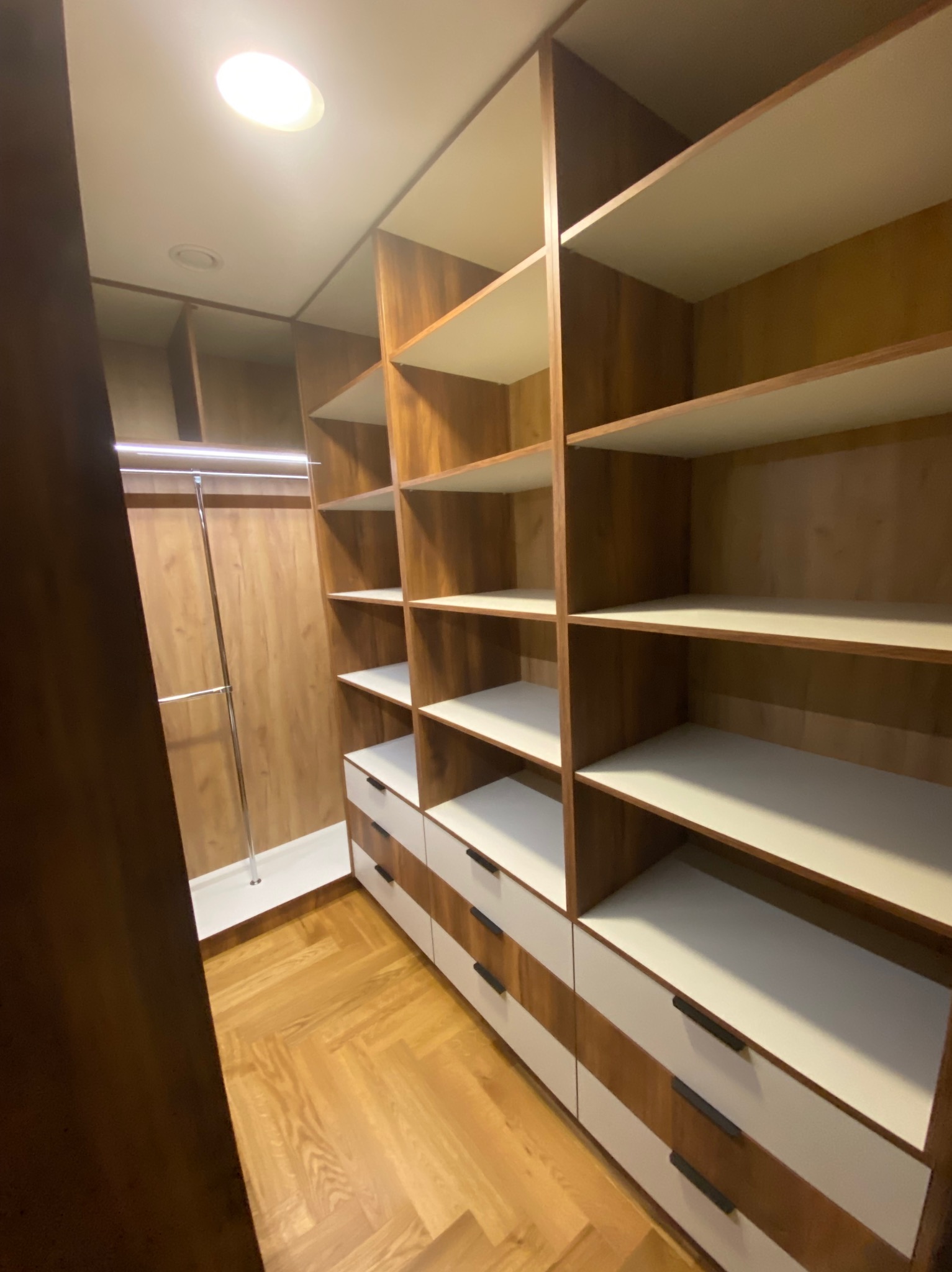 Wnętrze garderoby z drewnianymi półkami i białymi szufladami, oświetlona lampą sufitową. Widoczny parkiet ułożony w jodełkę i drążek na ubrania.