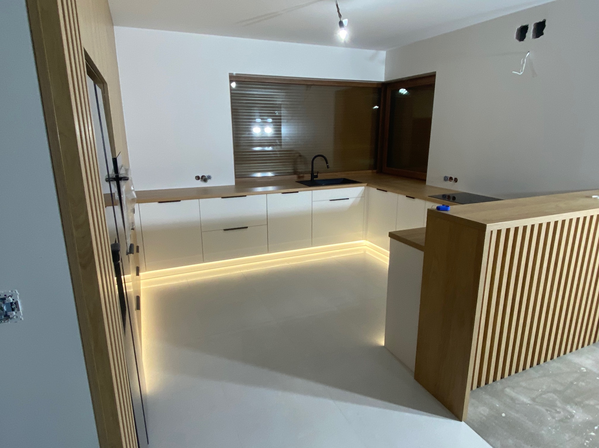 Nowoczesna kuchnia z białymi szafkami, drewnianym blatem i oświetleniem LED pod szafkami. Widoczny fragment wyspy kuchennej z drewnianymi lamelami.