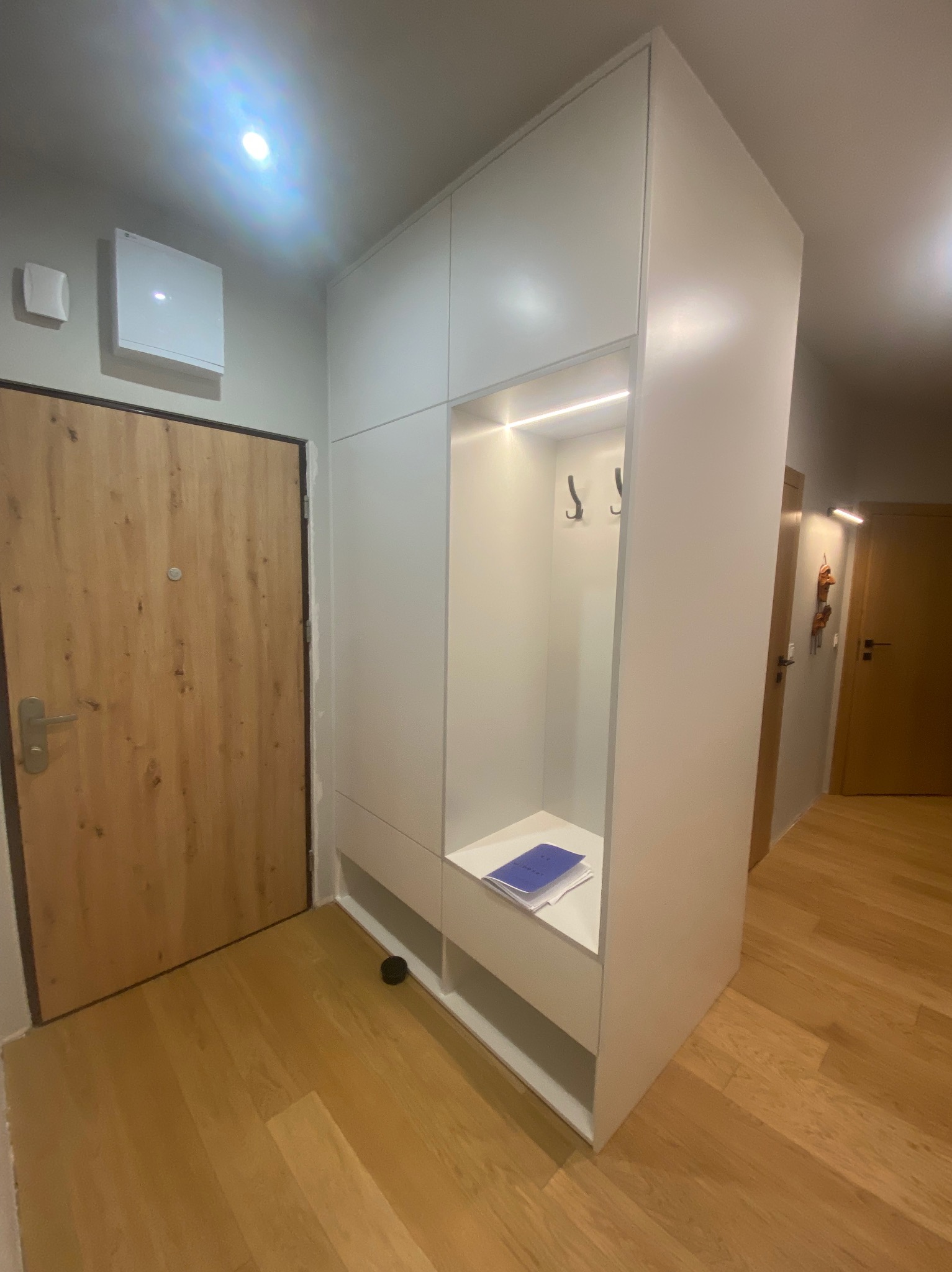 Biała szafa wnękowa z otwartą przestrzenią na buty i wieszaki, oświetlona LED, w przedpokoju z drewnianą podłogą i drzwiami.