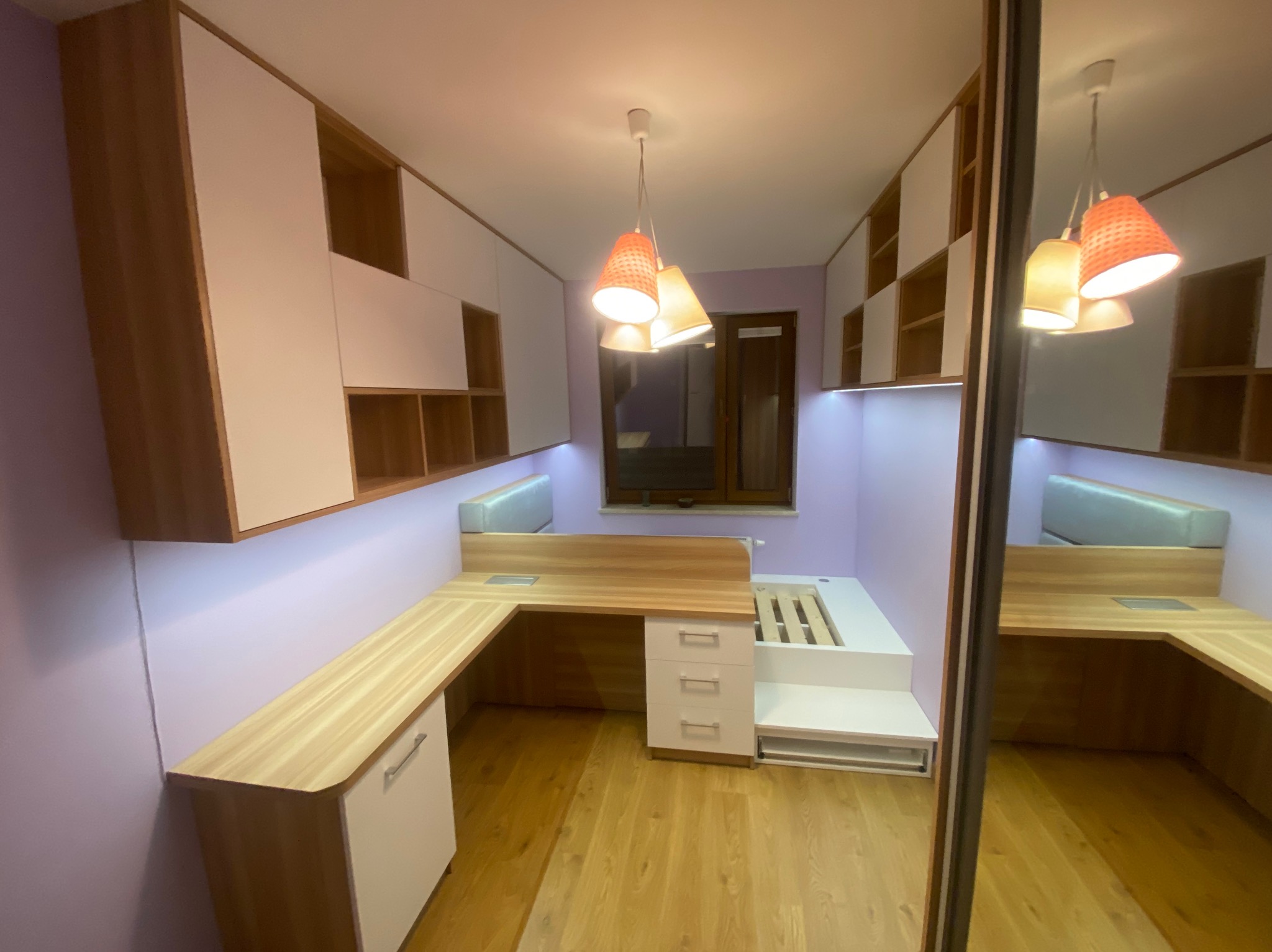 Pokój młodzieżowy z zabudową: biurko z szafkami, szafki wiszące z otwartymi półkami i łóżko na podeście. Całość w jasnym drewnie i bieli, oświetlenie punktowe.