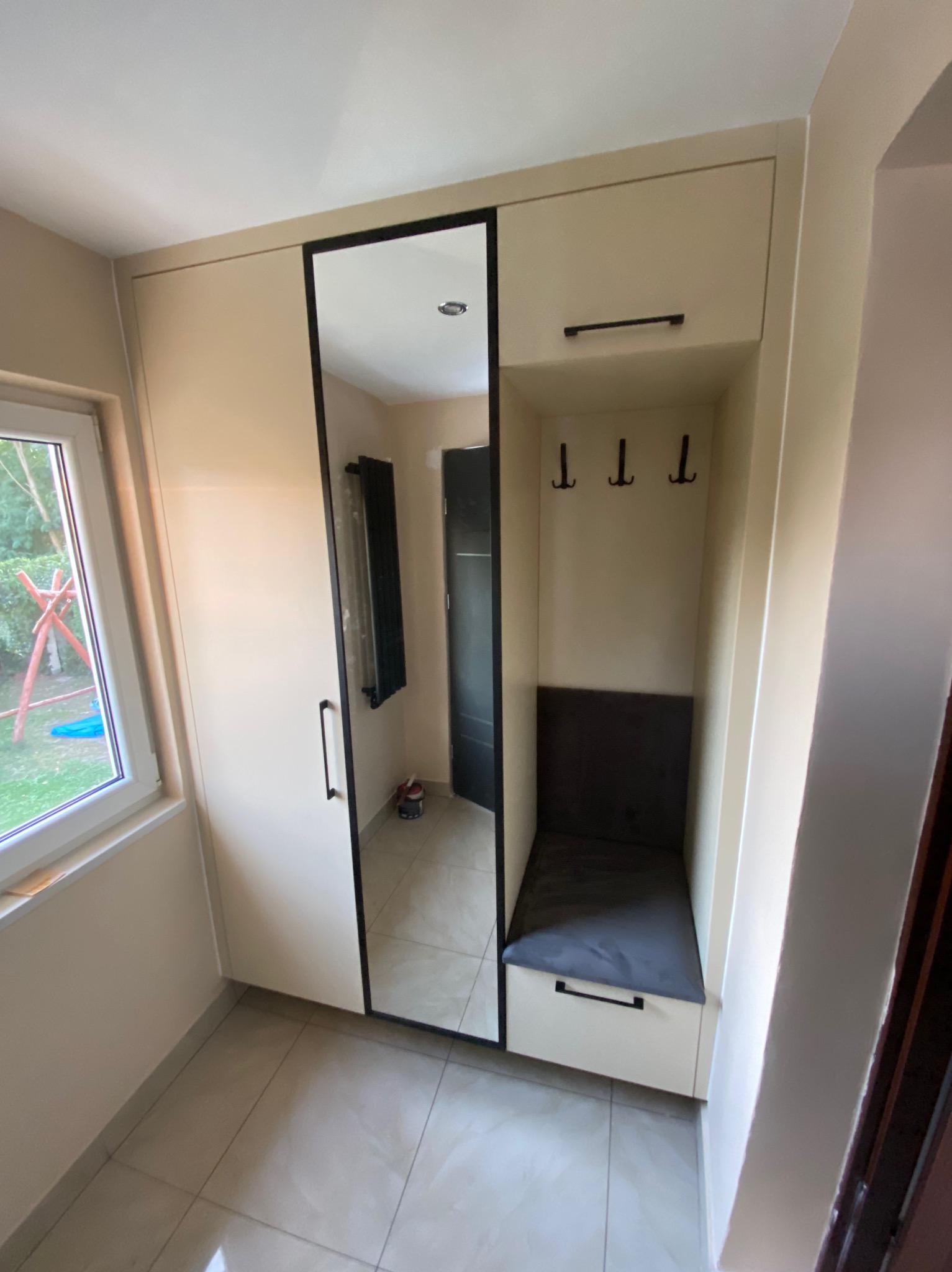 Beżowa szafa wnękowa z lustrem w czarnej ramie i siedziskiem z szufladą, uzupełniona o wieszaki. Widok na wnętrze przez lustro.