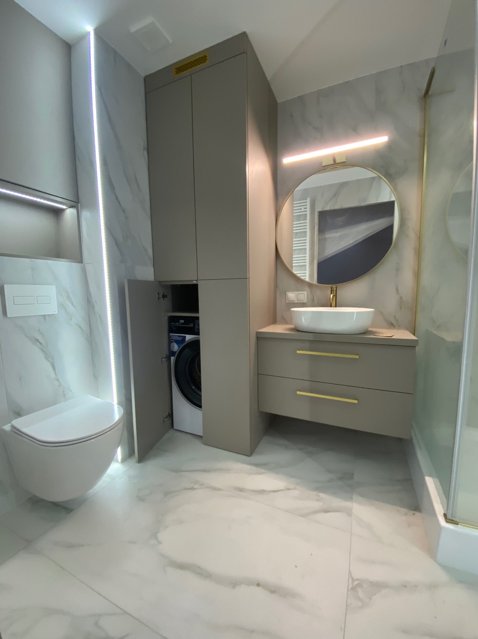 Nowoczesna łazienka z zabudową na pralkę, umywalką nablatową i okrągłym lustrem z podświetleniem LED. Minimalistyczny design, marmurowe płytki i złote akcenty.