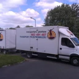 Biały samochód dostawczy z przyczepą kontenerową firmy z Piotrkowa Trybunalskiego, z widocznym logo i numerem telefonu, zaparkowany na asfalcie w otoczeniu zieleni.