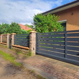 Konstrukcje Metalowe Marcin Szczesny - Nowoczesna, przesuwna brama wjazdowa z poziomymi panelami w kolorze antracytu, osadzona między kamiennymi słupkami. W tle widoczny fragment domu i ogrodzenie.