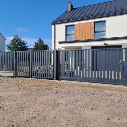 Konstrukcje Metalowe Marcin Szczesny - Nowoczesna, przesuwna brama wjazdowa z metalowymi sztachetami w kolorze grafitowym, na tle jasnego domu z ciemnym dachem i błękitnego nieba. Widoczny fragment posesji.