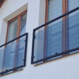 Konstrukcje Metalowe Marcin Szczesny - Dwa balkony z nowoczesnymi, szklanymi balustradami w czarnych, metalowych ramach, zamontowane na białej fasadzie budynku z drewnianymi oknami.