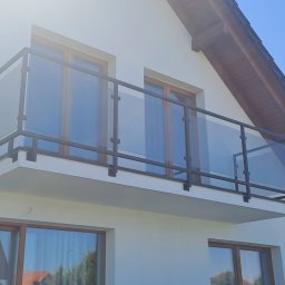 Konstrukcje Metalowe Marcin Szczesny - Nowoczesna balustrada balkonowa z czarną metalową ramą i przezroczystym szkłem, zamontowana na białym budynku z drewnianymi oknami. Widok z dołu, błękitne niebo w tle.