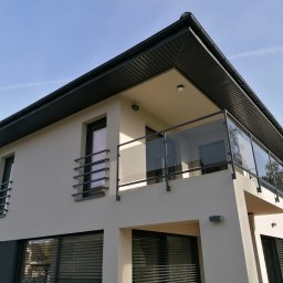 Konstrukcje Metalowe Marcin Szczesny - Nowoczesny dom z balkonem zabezpieczonym balustradą ze szkła i metalu. Minimalistyczny design, jasna elewacja, ciemne okna i dach. Ujęcie z dołu, błękitne niebo w tle.