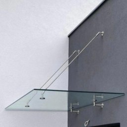 Konstrukcje Metalowe Marcin Szczesny - Szklany daszek nad wejściem, wsparty na metalowych linkach i uchwytach, minimalistyczny design, szara ściana budynku.