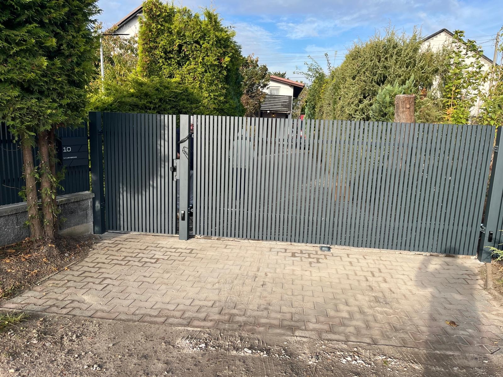 Nowoczesna, szara brama wjazdowa z pionowych profili, częściowo otwarta, z widocznym numerem domu i brukowanym podjazdem. Tło stanowią drzewa i fragment domu.