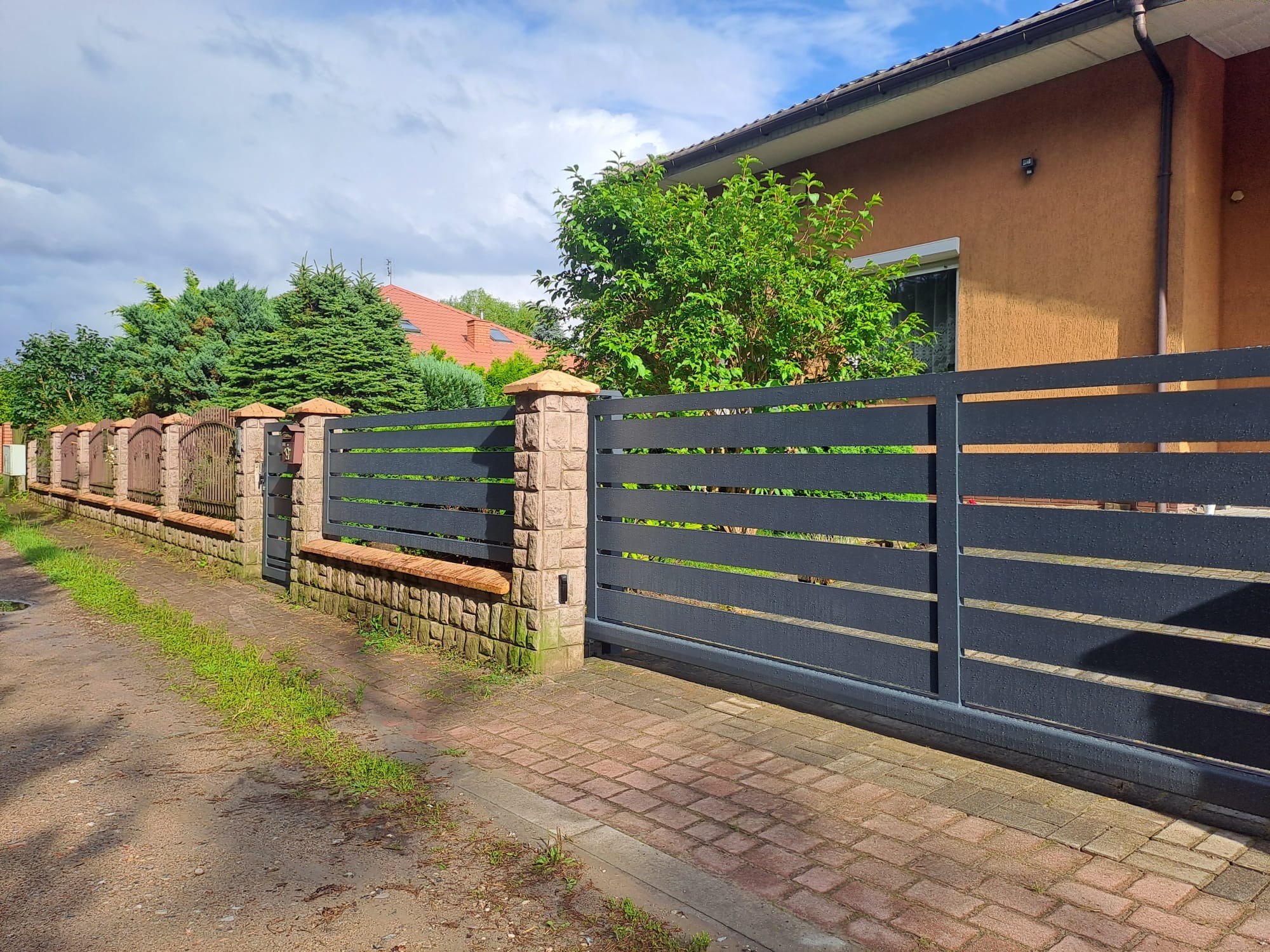 Nowoczesna, przesuwna brama wjazdowa z poziomymi panelami w kolorze antracytu, osadzona między kamiennymi słupkami. W tle widoczny fragment domu i ogrodzenie.