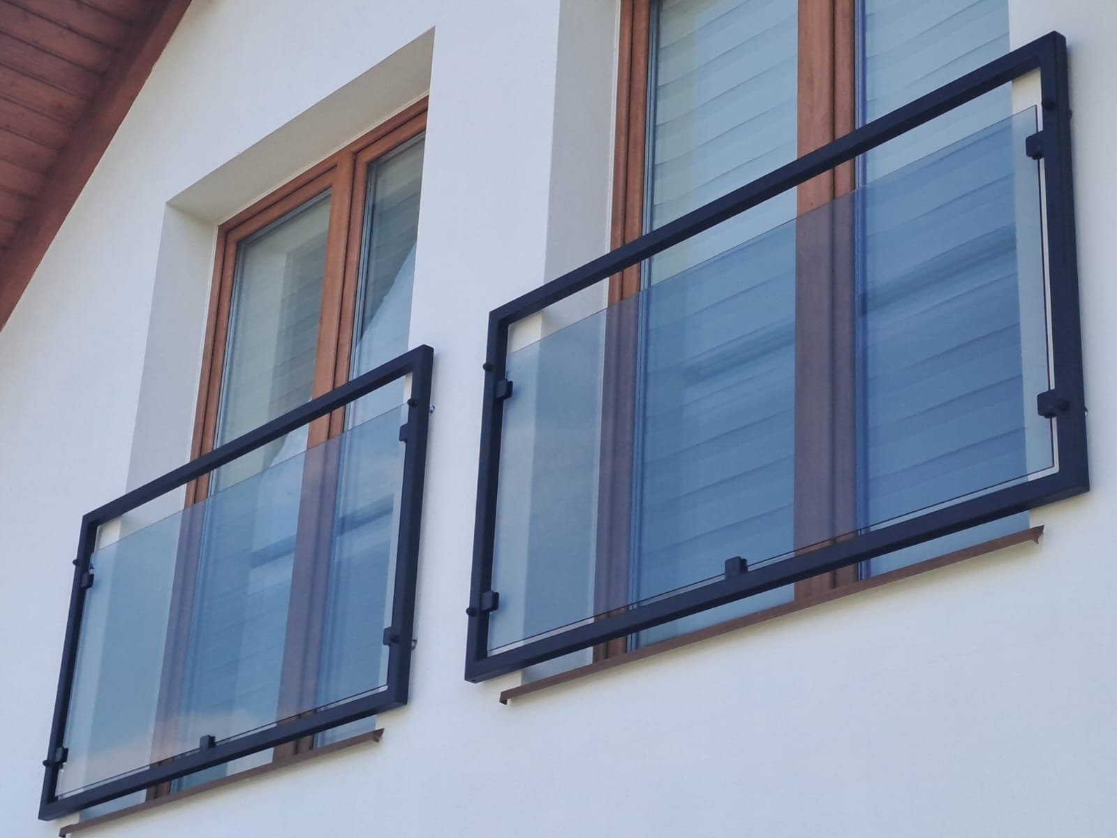 Dwa balkony z nowoczesnymi, szklanymi balustradami w czarnych, metalowych ramach, zamontowane na białej fasadzie budynku z drewnianymi oknami.
