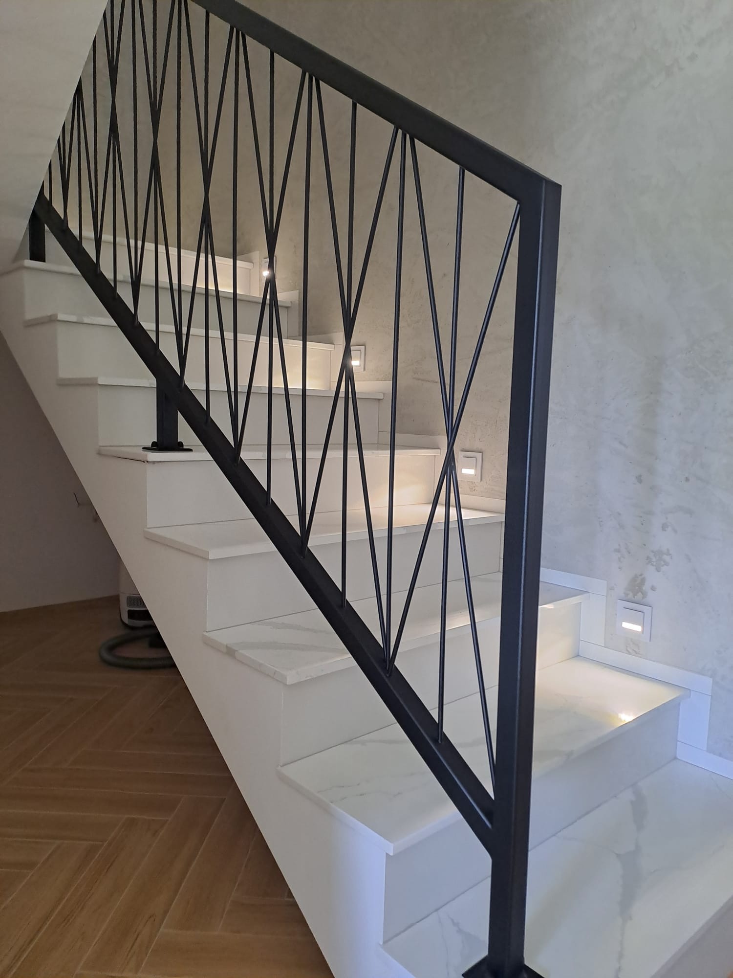 Nowoczesne schody z białą okładziną i czarną, geometryczną balustradą. Oświetlenie LED w ścianie i na stopniach. Parkiet w jodełkę na podłodze.