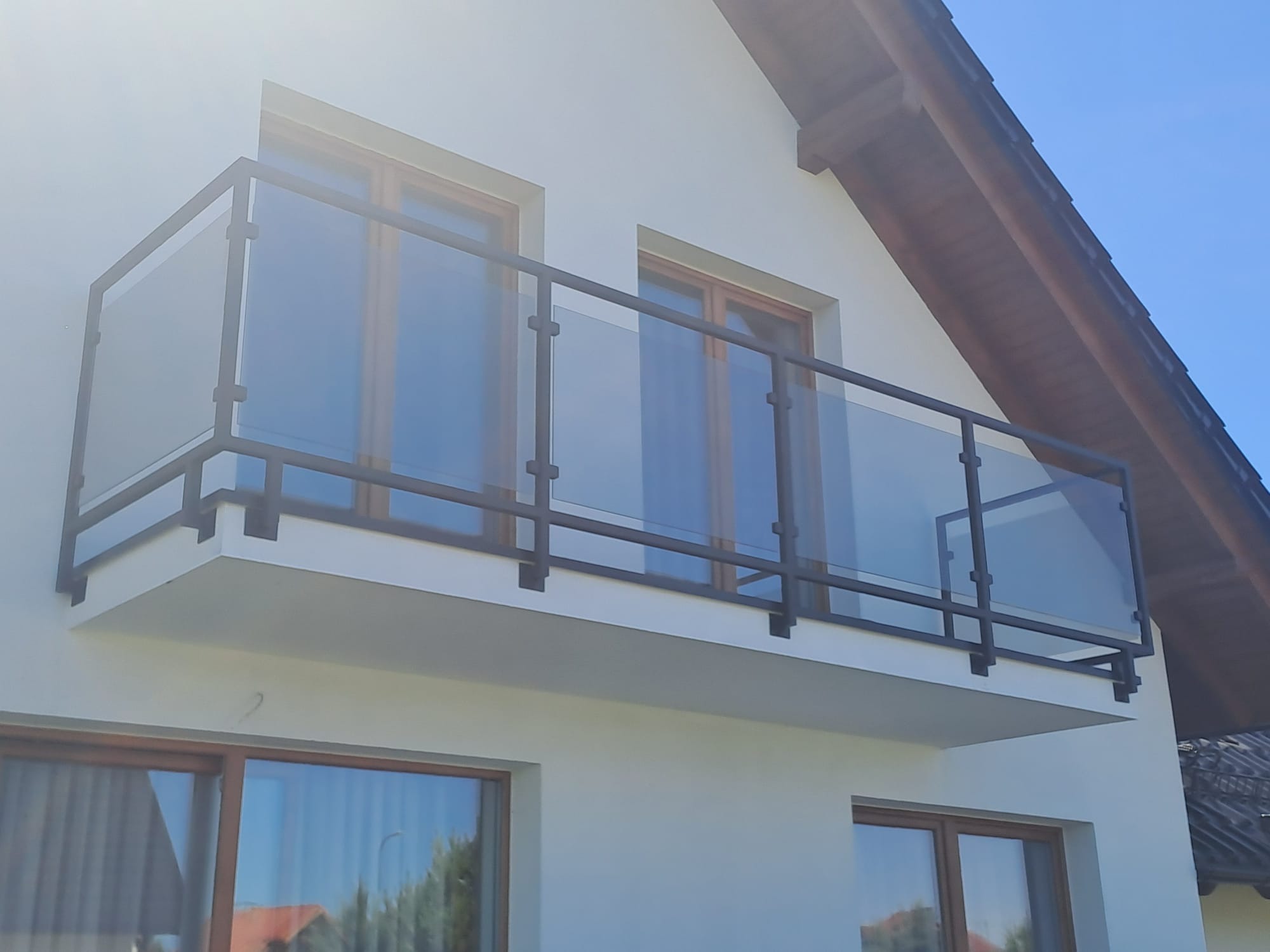Nowoczesna balustrada balkonowa z czarną metalową ramą i przezroczystym szkłem, zamontowana na białym budynku z drewnianymi oknami. Widok z dołu, błękitne niebo w tle.