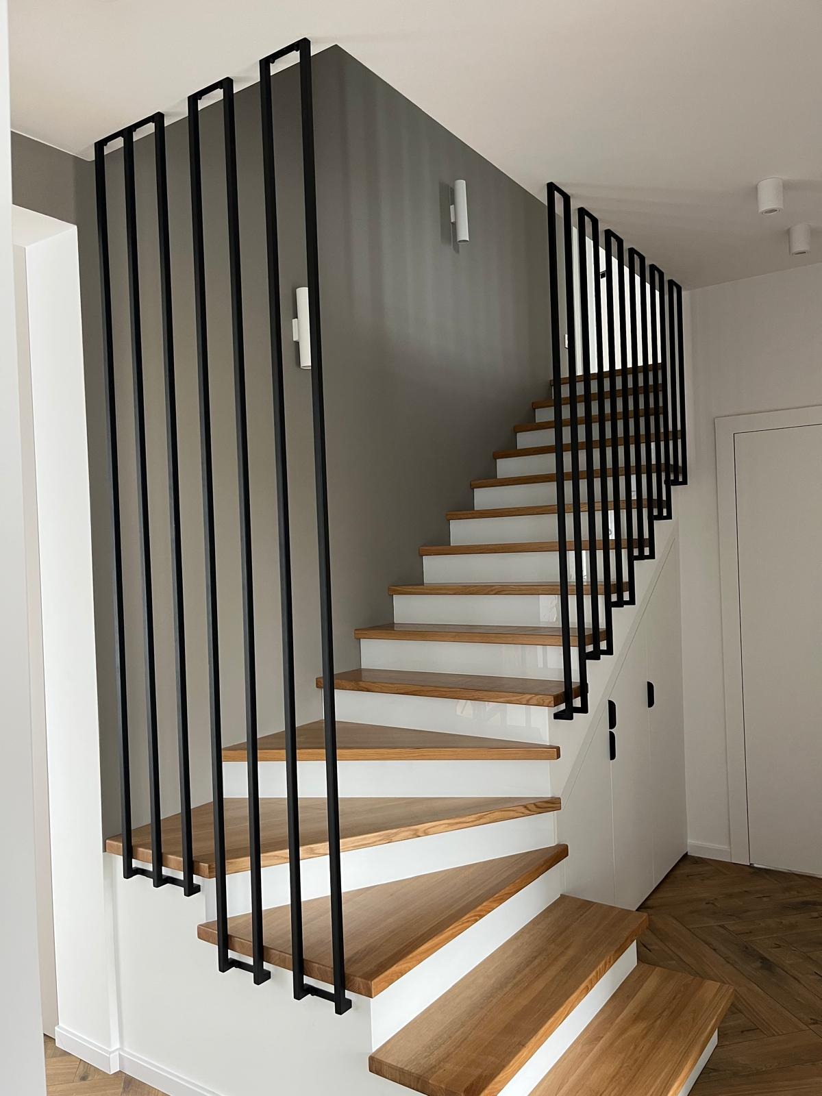 Nowoczesne schody drewniane z czarną, minimalistyczną balustradą z pionowych prętów. Jasne wnętrze, parkiet w jodełkę. Ujęcie z dołu, widok na bieg schodów.