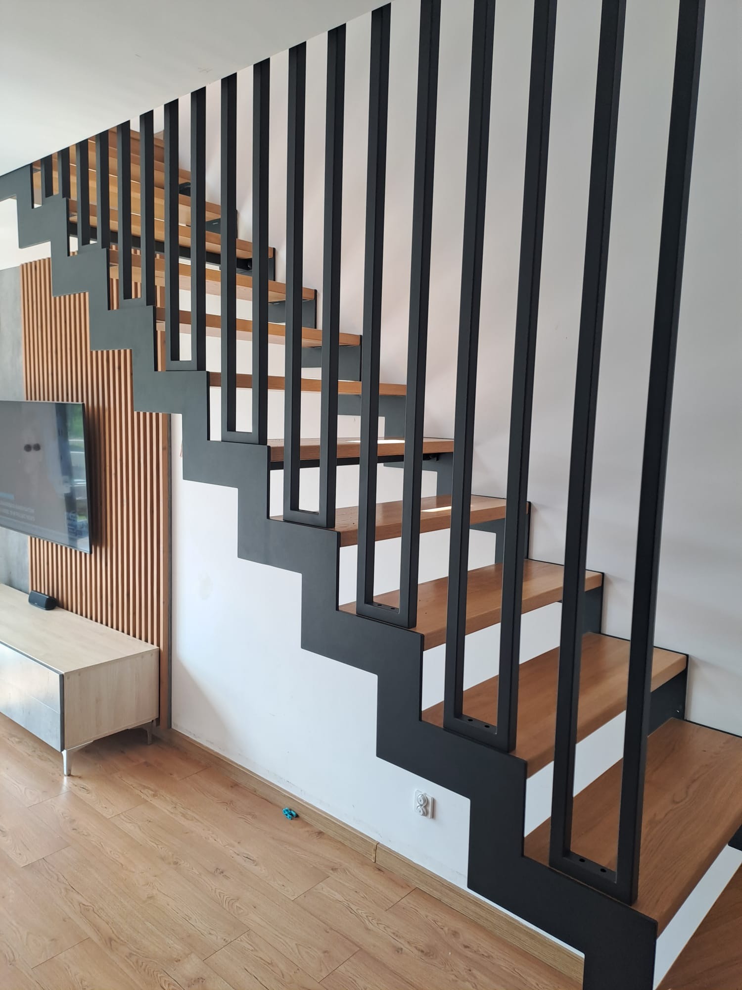 Nowoczesne schody z drewnianymi stopniami i czarną, minimalistyczną balustradą z pionowych prętów. Widok z dołu, jasne wnętrze.