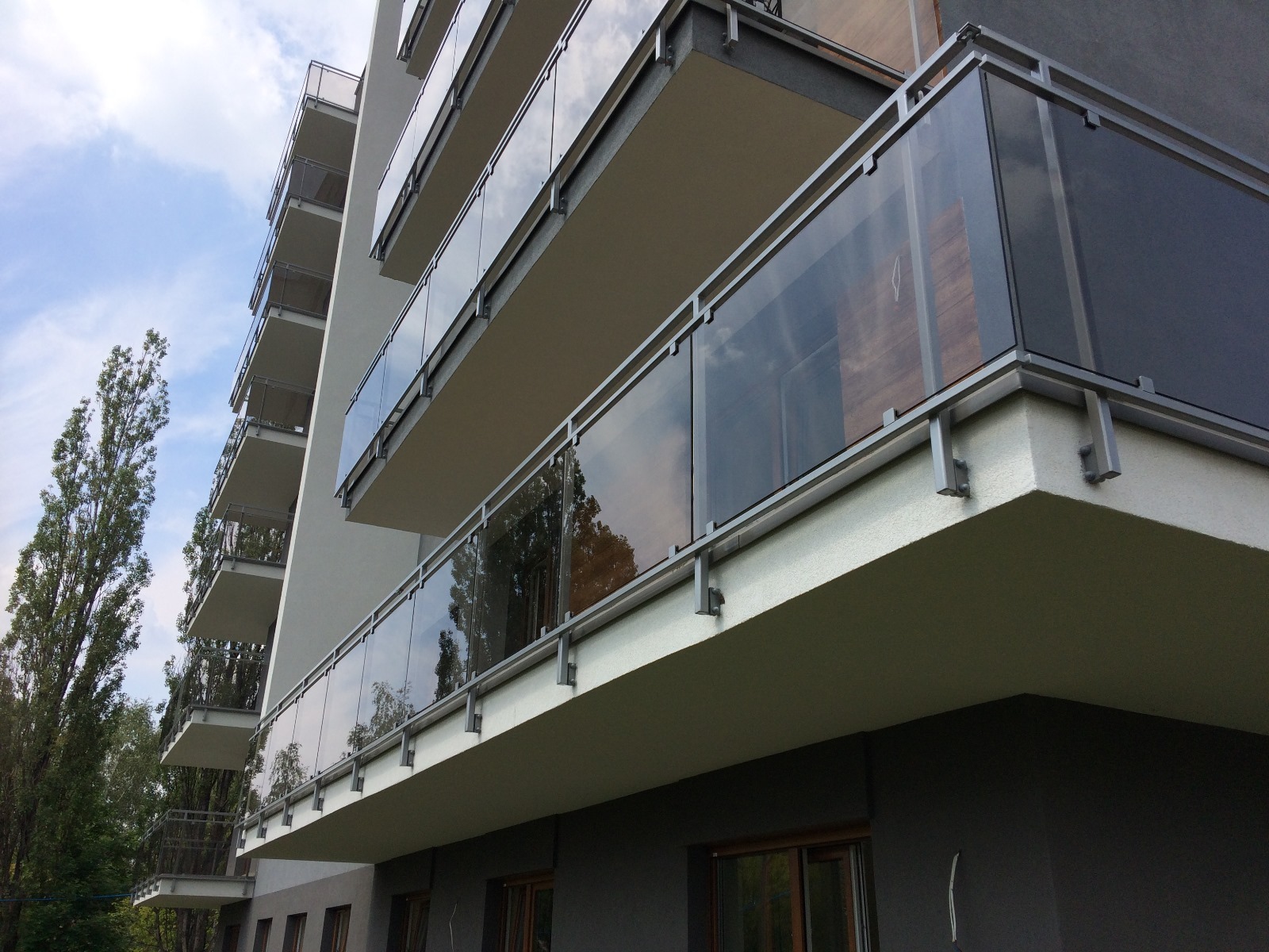 Nowoczesne, szklane balustrady balkonowe z metalowymi ramami, zamontowane w budynku wielorodzinnym. Widok z dołu, podkreślający minimalistyczny design i przejrzystość konstrukcji.