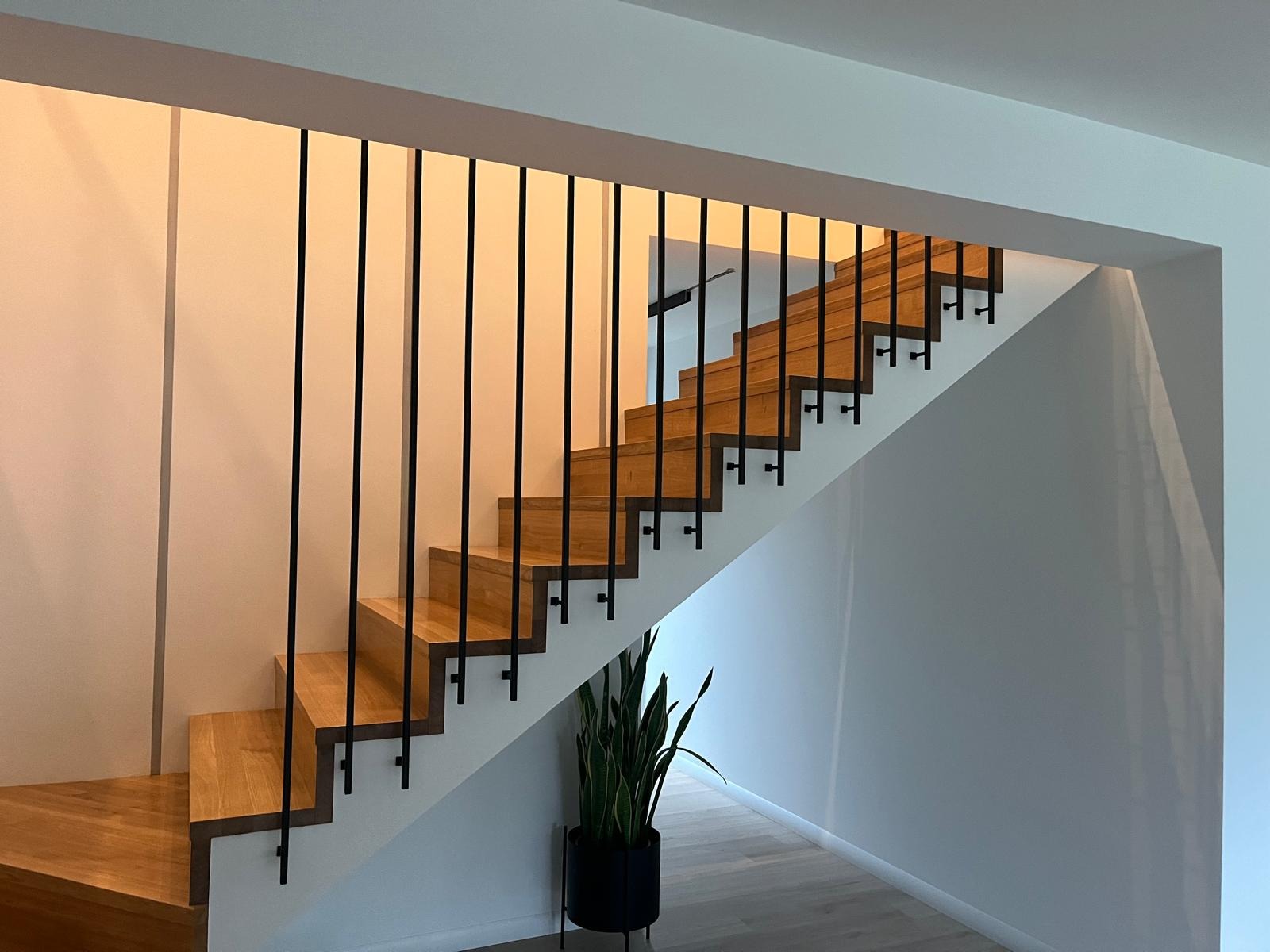 Drewniane schody z czarnymi, minimalistycznymi poręczami w nowoczesnym wnętrzu. U dołu widoczna roślina doniczkowa. Jasne ściany i podłoga.