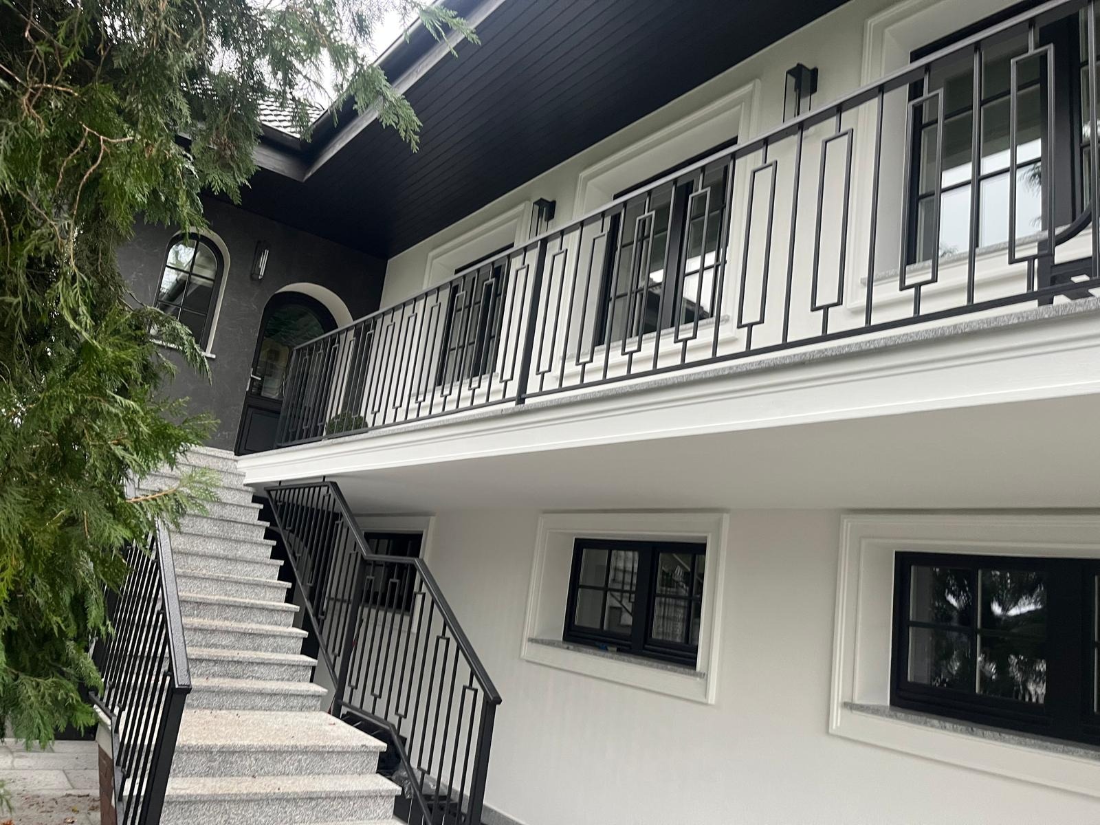 Eleganckie schody zewnętrzne z szarego granitu prowadzą na balkon z czarną, geometryczną balustradą. Biała fasada budynku z czarnymi oknami i dachem.