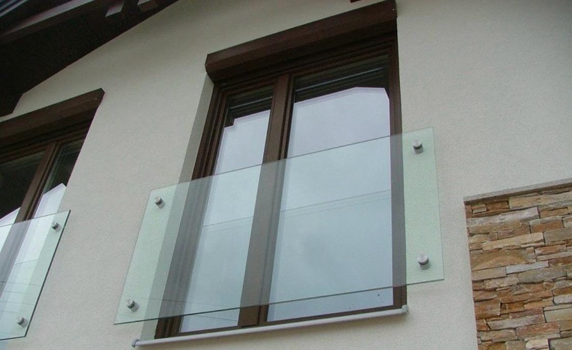 Szklana balustrada balkonowa z brązowymi ramami okiennymi i roletami zewnętrznymi. Widok z dołu na elewację budynku z kamiennym wykończeniem.