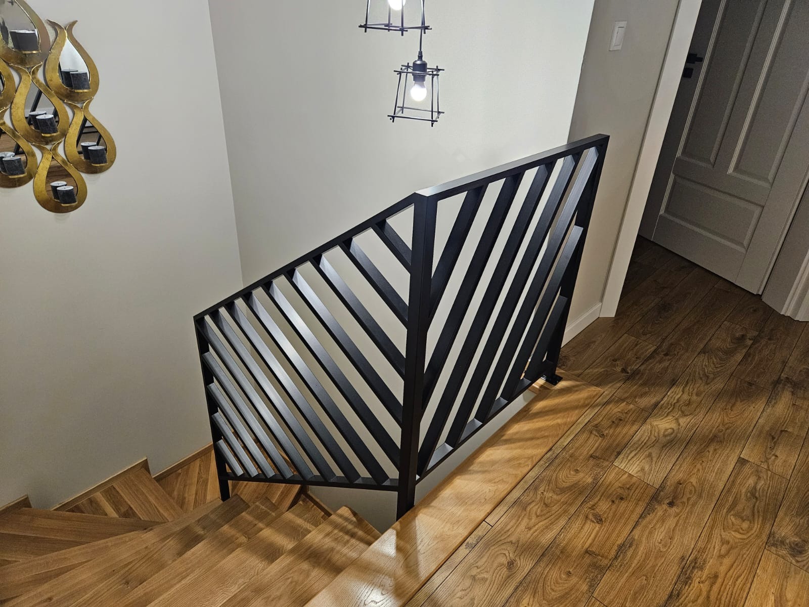 Nowoczesna, czarna balustrada schodowa z ukośnymi prętami, widziana z góry. Drewniane schody i podłoga, minimalistyczne oświetlenie. Dekoracyjne lustro na ścianie.