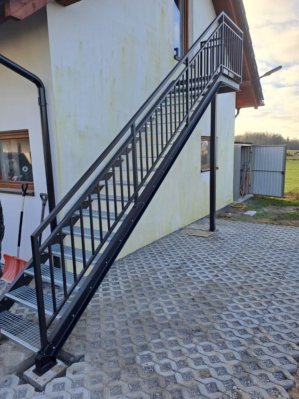Zewnętrzne schody metalowe z czarną balustradą prowadzące do drzwi na piętrze, z widoczną kratką na stopniach i kostką ażurową na podłożu.