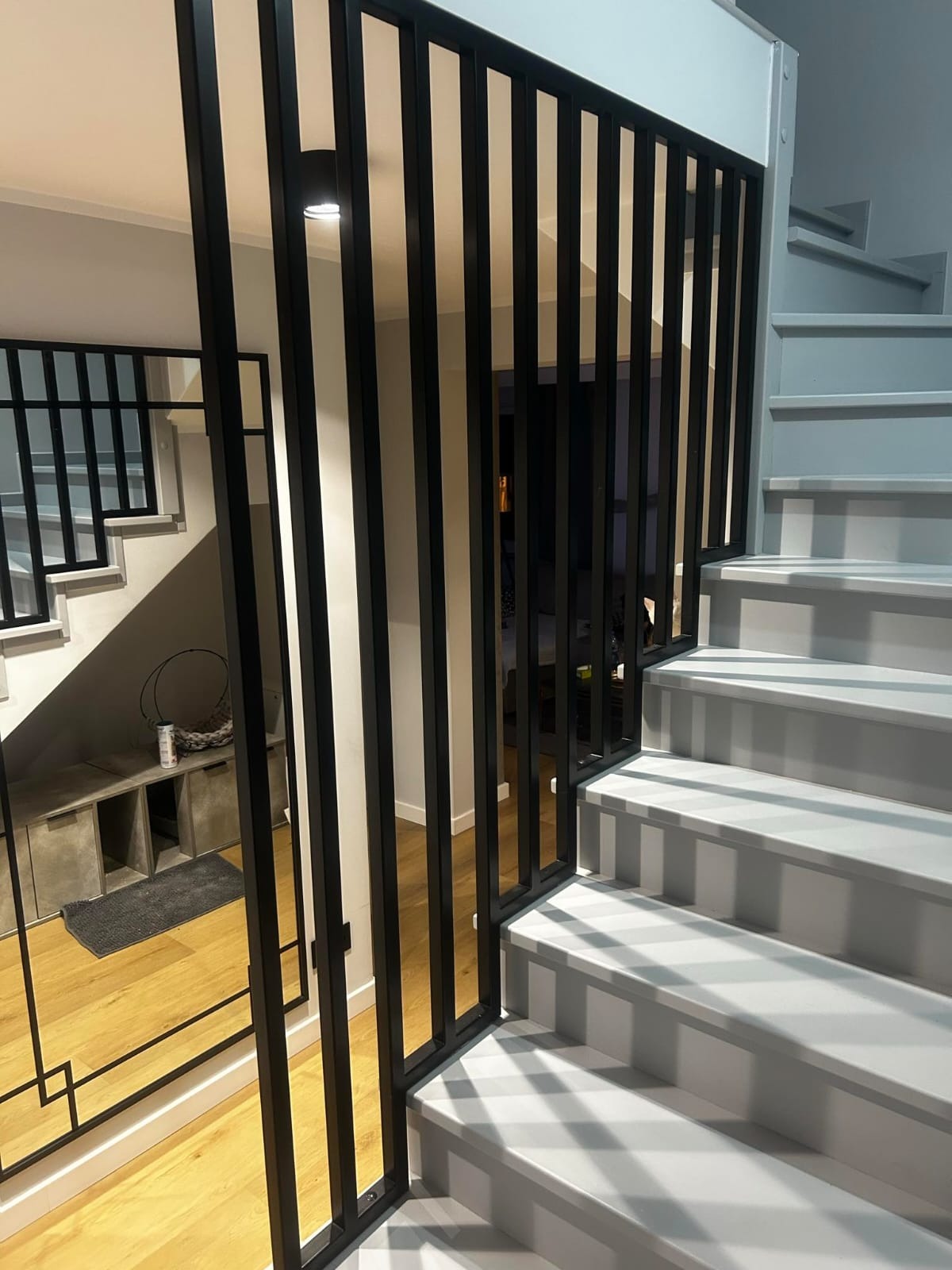 Nowoczesne schody z czarną, pionową balustradą. Białe stopnie schodów kontrastują z ciemną balustradą. Widoczny fragment podłogi i schody w tle.