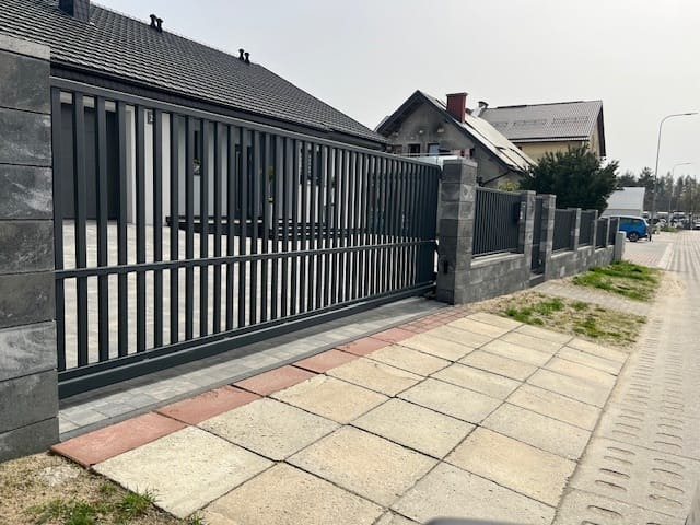Nowoczesna, przesuwna brama wjazdowa z metalowymi sztachetami, osadzona na betonowych słupkach. Widok z zewnątrz posesji, z kostką brukową na pierwszym planie.