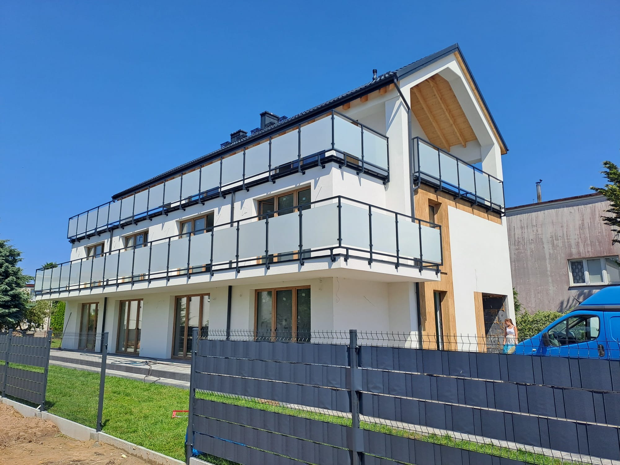 Nowoczesny dom z balkonami zabezpieczonymi szklanymi balustradami i czarnymi poręczami. W tle błękitne niebo i fragment ogrodzenia panelowego.