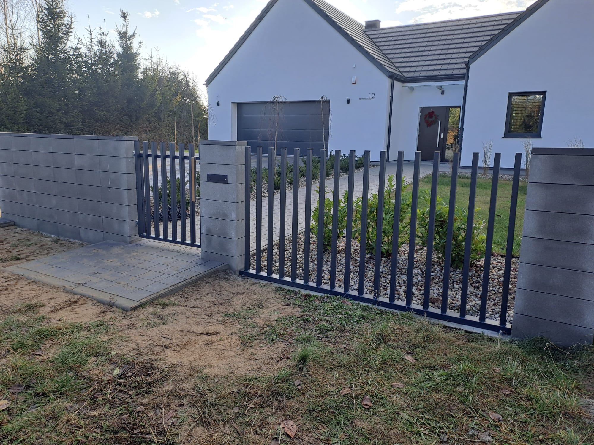 Nowoczesne ogrodzenie posesji z metalowymi przęsłami i betonowymi słupkami, prowadzące do domu z szarą bramą garażową i numerem 12. Widok z zewnątrz.