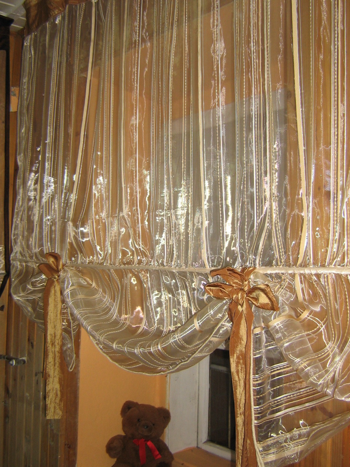 Przezroczysta, marszczona firanka z kokardami w złotym odcieniu, wisząca w oknie, z pluszowym misiem w rogu kadru.