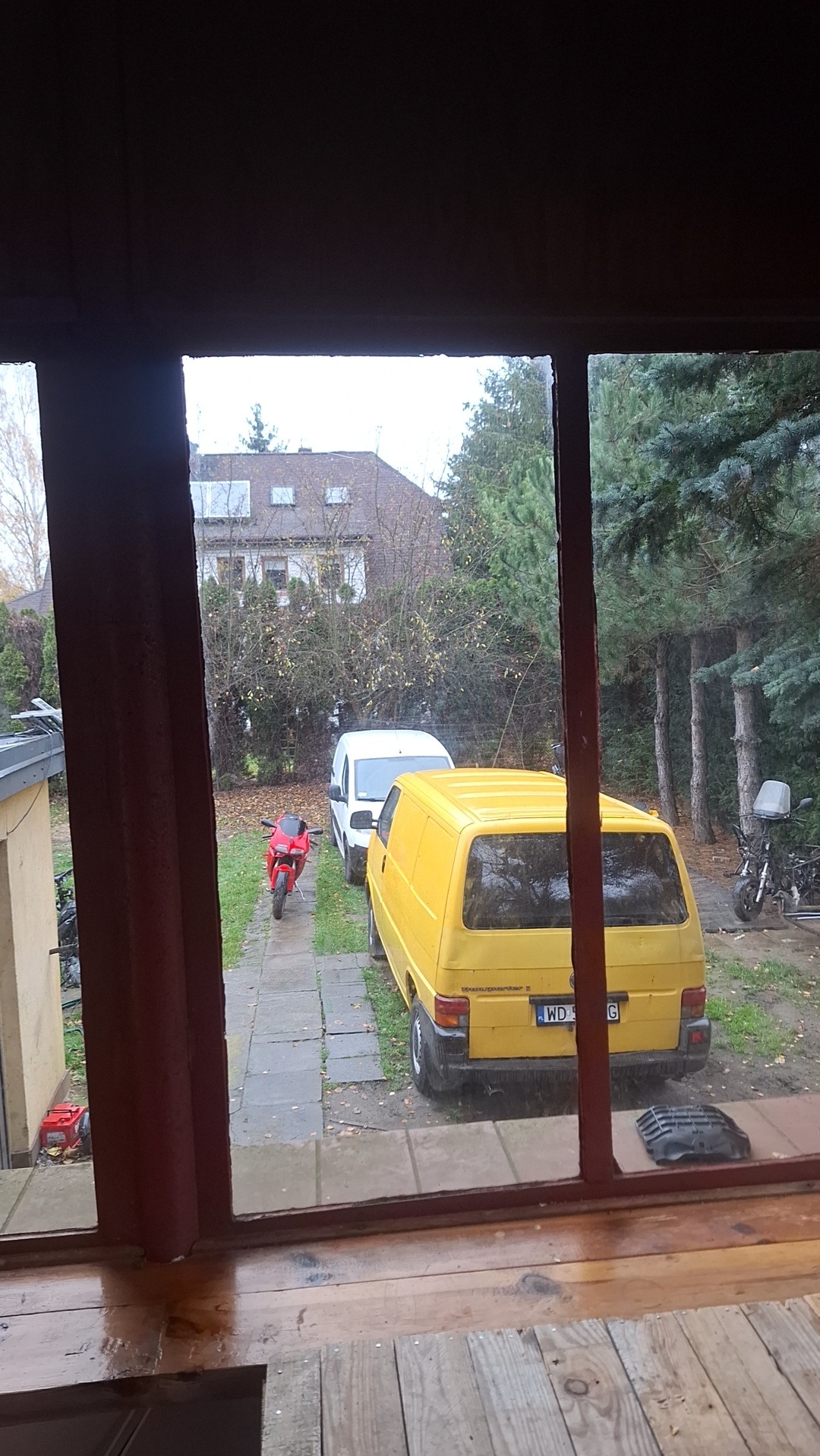 Żółty bus VW Transporter zaparkowany na podwórku, widziany przez okno. Obok biały bus i czerwony motocykl. W tle dom i drzewa. Pochmurny dzień.