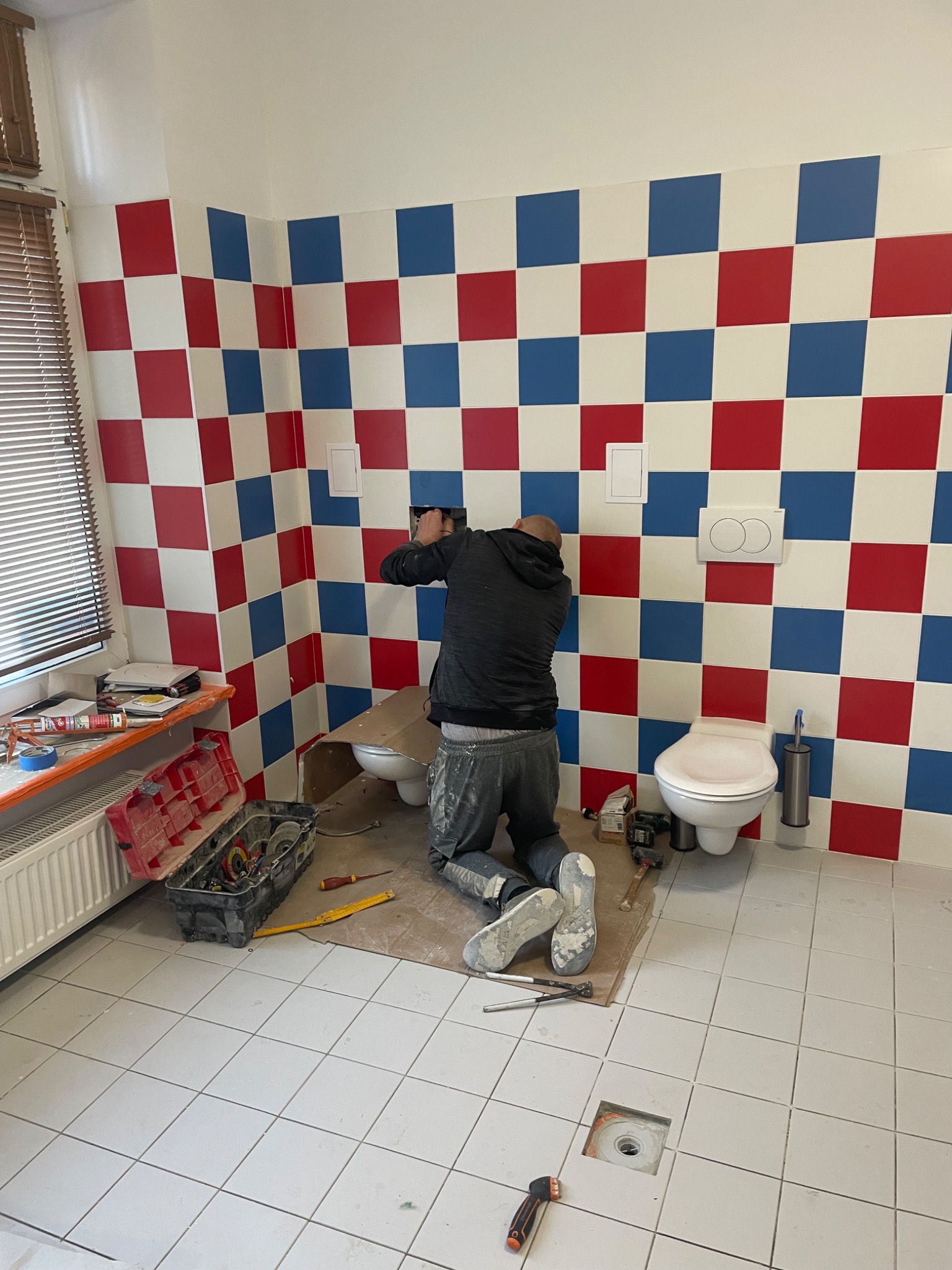 Instalator w trakcie montażu armatury w łazience z białą, kwadratową podłogą i ścianami w biało-czerwono-niebieską szachownicę. Widoczne narzędzia i elementy instalacji.