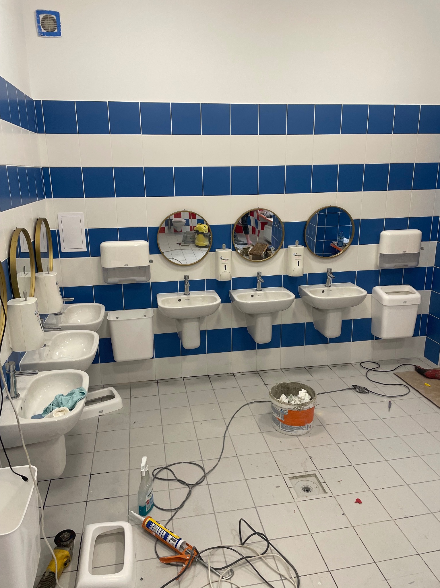 Wykończona łazienka w stylu marynistycznym z trzema umywalkami i okrągłymi lustrami. Widoczne narzędzia i akcesoria, sugerujące świeżo zakończone prace wykończeniowe.