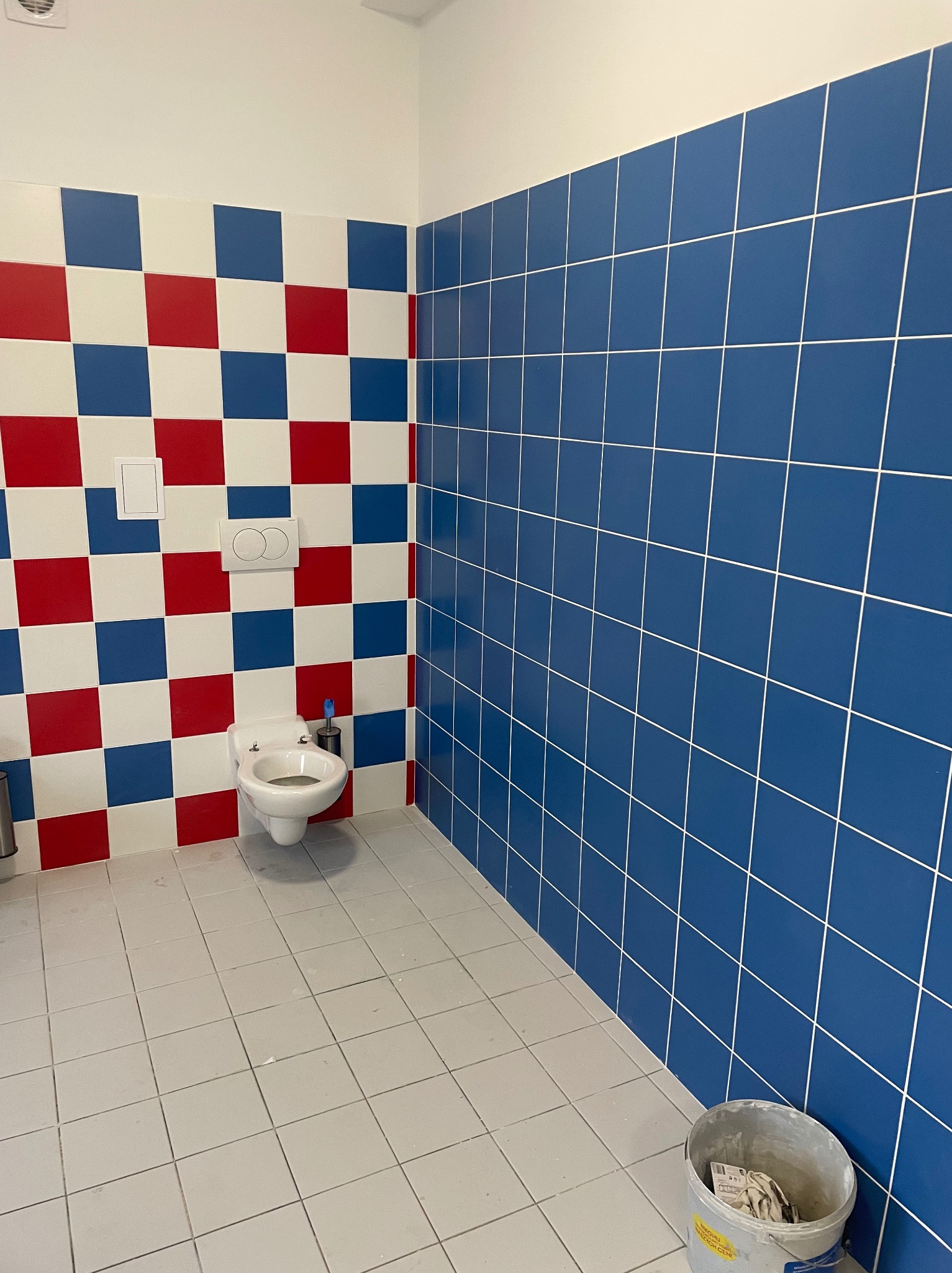 Wykończona łazienka z toaletą, ściany w niebieskie płytki i szachownicę czerwono-biało-niebieską. Na podłodze szare płytki. Widoczne wiadro po farbie.