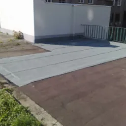 Płaski dach pokryty szarą papą termozgrzewalną z widocznymi łączeniami, zabezpieczony zieloną barierką, przylegający do białego budynku.