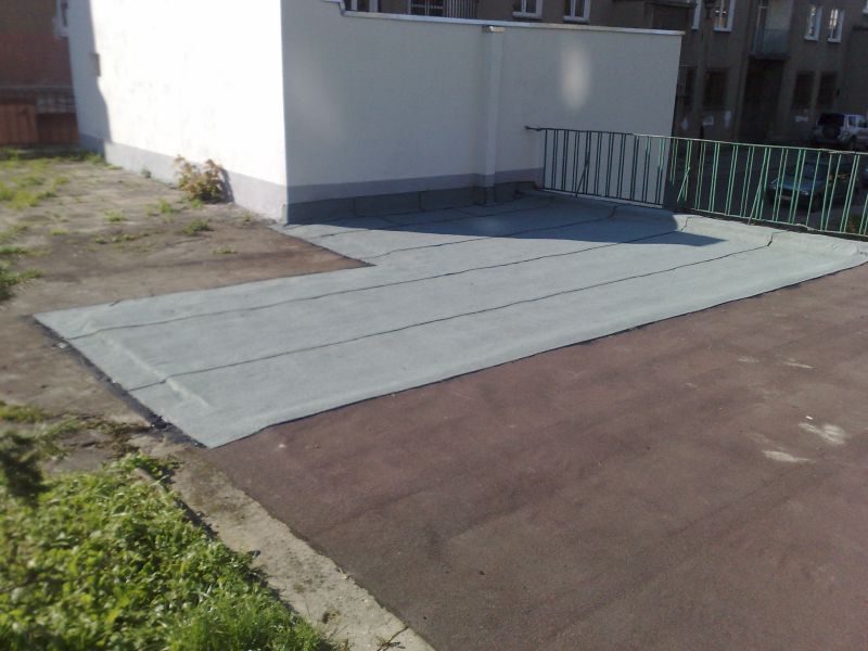 Płaski dach pokryty szarą papą termozgrzewalną z widocznymi łączeniami, zabezpieczony zieloną barierką, przylegający do białego budynku.