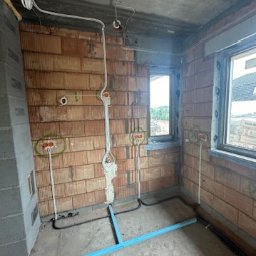 Elopro - Surowy mur z widocznymi przewodami elektrycznymi i puszkami instalacyjnymi. Widoczne okno i fragment podłogi z niebieskimi profilami.