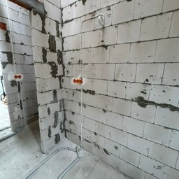 Elopro - Surowe ściany z betonu komórkowego z widocznymi puszkami elektrycznymi i przewodami w peszlach, ułożonymi na podłodze. W tle widoczne przejście do innego pomieszczenia.