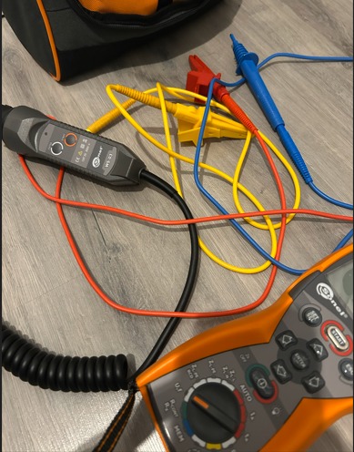 Miernik elektryczny z przewodami i torbą narzędziową na podłodze. Widoczne kable w kolorach: czerwonym, żółtym i niebieskim. Sprzęt pomiarowy marki Sonel.