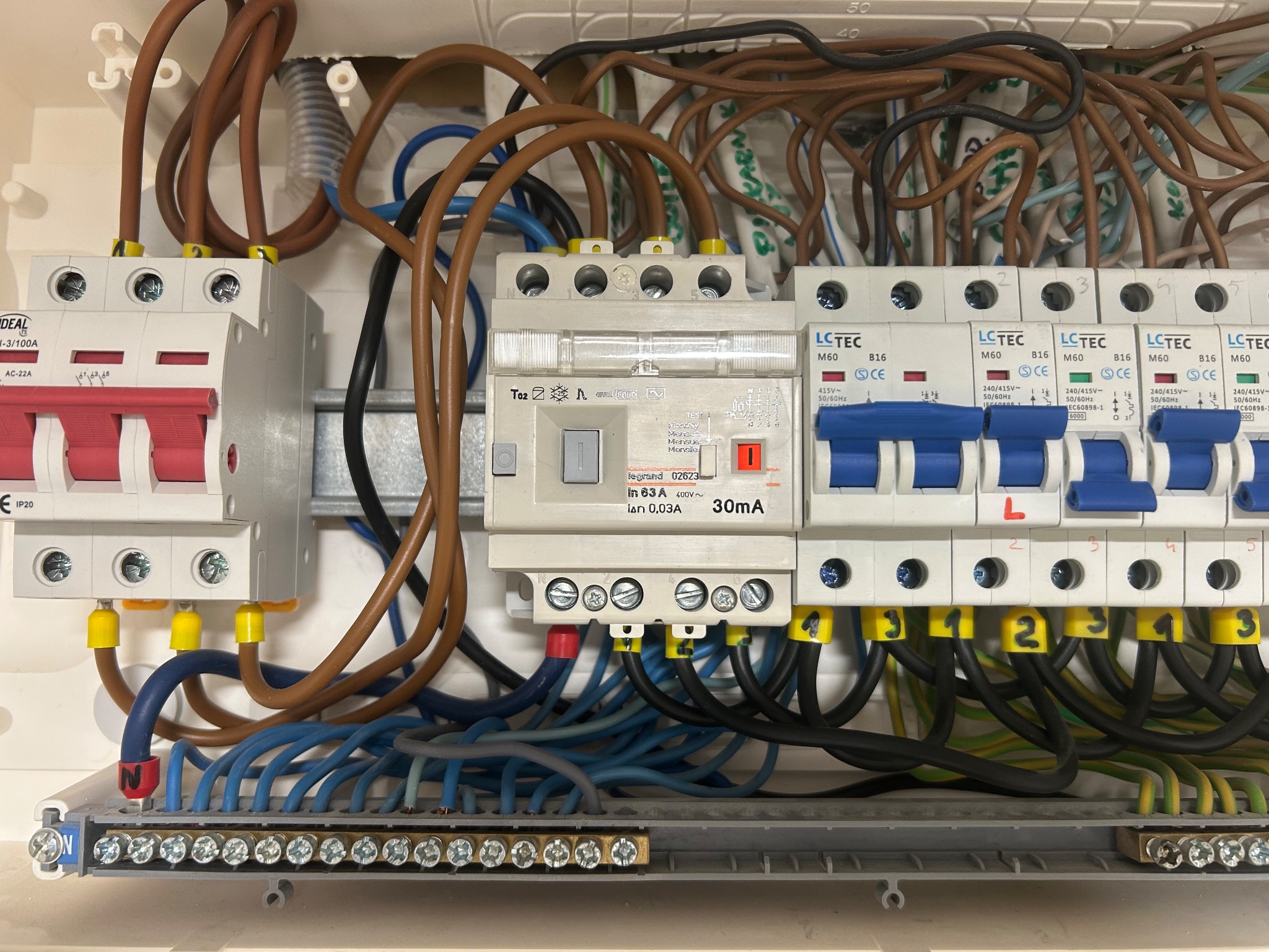 Otwarta skrzynka elektryczna z widocznymi bezpiecznikami, przewodami o różnych kolorach i oznaczeniami. Widoczne żółte oznaczenia na końcówkach przewodów.