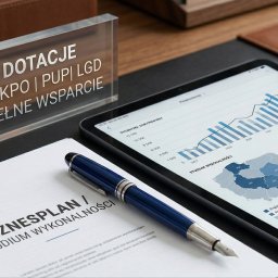 Kamil Trela - Stół z tabletem prezentującym wykresy, dokumentem z napisem 'Biznesplan' i akrylową tabliczką 'Dotacje UE KPO'. Eleganckie pióro obok.