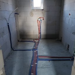 Aqua - Instalacja wodna z rur PEX w surowym wnętrzu. Czerwone i niebieskie rury prowadzone wzdłuż ścian i podłogi, zabezpieczone uchwytami. Widoczne przyłącza i okno.