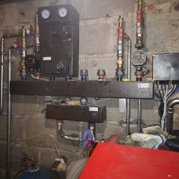 Aqua - Rozbudowana instalacja CO z czarnym rozdzielaczem Afriso na tle surowej ściany z betonu komórkowego. Widoczne zawory, pompy obiegowe i manometry.