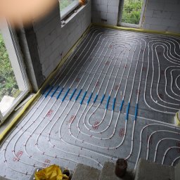 Aqua - Białe rury ogrzewania podłogowego z niebieskimi łącznikami, ułożone na szarej macie izolacyjnej w surowym wnętrzu z oknami. Widoczne elementy montażowe i narzędzia.