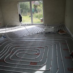 Aqua - Białe rury ogrzewania podłogowego ułożone na szarej macie z czerwonymi elementami mocującymi, widok z góry na instalację w surowym wnętrzu z oknem.