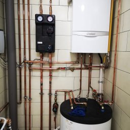 Aqua - Złożona instalacja CO z piecem gazowym Vaillant, zbiornikiem i czarnym modułem sterującym, rury miedziane prowadzone wzdłuż kafelkowanej ściany. Widok na kotłownię.